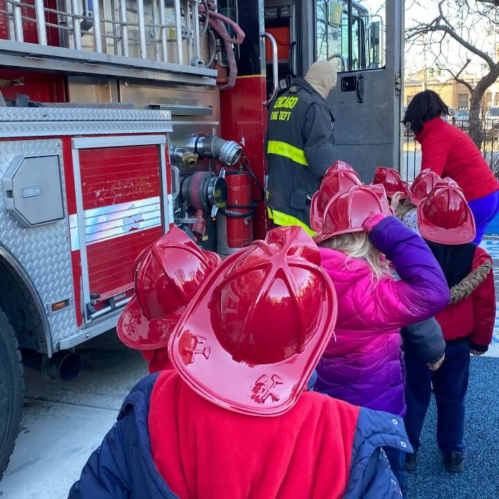 Children wearing plastic firefighter helmets line up near a fire truck.
